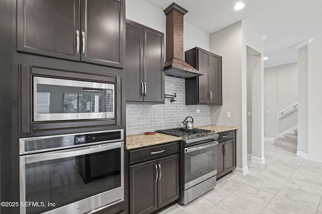 kitchen with premium range hood, appliances with stainless steel finishes, backsplash, dark brown cabinets, and light stone counters