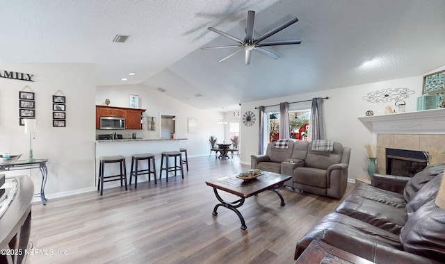 living room featuring lofted ceiling, a tiled fireplace, ceiling fan, a textured ceiling, and light hardwood / wood-style flooring