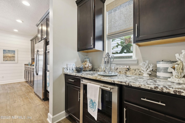 kitchen with dark brown cabinetry, stainless steel fridge, wooden walls, beverage cooler, and light stone countertops