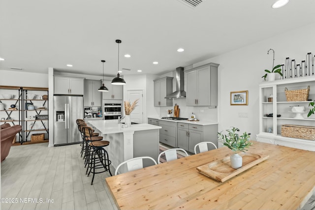 kitchen featuring light countertops, hanging light fixtures, appliances with stainless steel finishes, a kitchen island with sink, and wall chimney exhaust hood