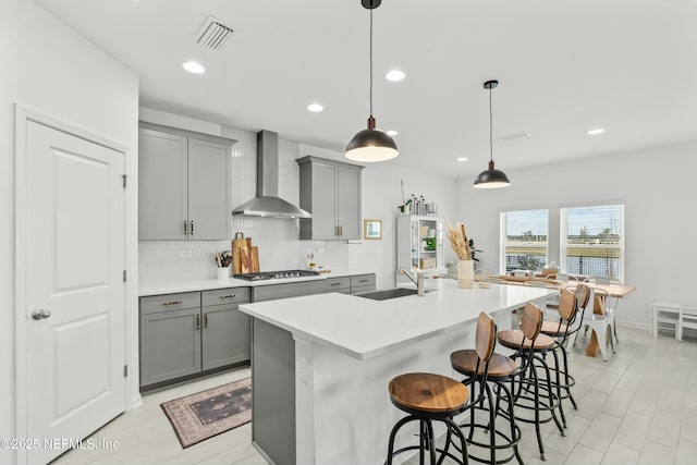 kitchen featuring a center island with sink, visible vents, light countertops, a sink, and wall chimney range hood