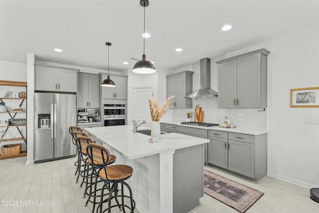 kitchen featuring stainless steel appliances, light countertops, a kitchen island with sink, a sink, and wall chimney range hood