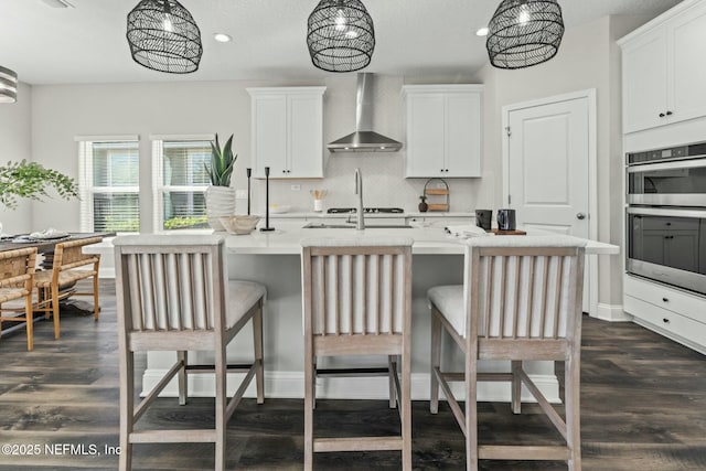 kitchen with white cabinets, stainless steel double oven, an island with sink, and wall chimney range hood