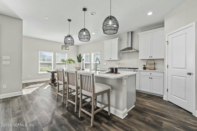 kitchen with sink, white cabinets, hanging light fixtures, wall chimney range hood, and a center island with sink