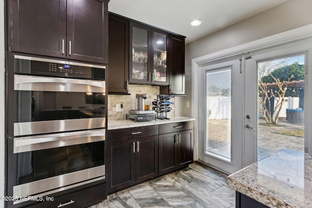 kitchen with dark brown cabinets, stainless steel double oven, light stone counters, and decorative backsplash