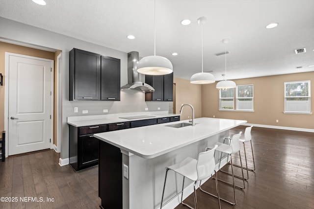 kitchen with sink, a center island with sink, pendant lighting, black electric stovetop, and wall chimney range hood