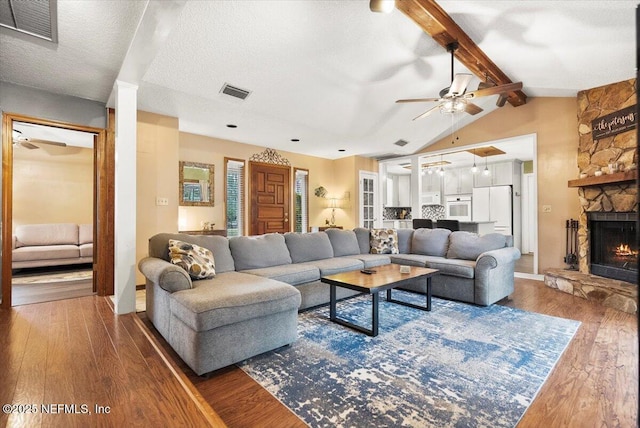 living area with ceiling fan, lofted ceiling with beams, a stone fireplace, visible vents, and dark wood-style floors