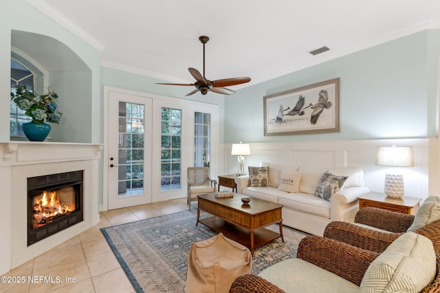 tiled living area featuring visible vents, ceiling fan, a wainscoted wall, ornamental molding, and a warm lit fireplace