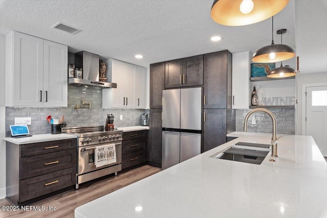 kitchen featuring wall chimney range hood, sink, white cabinetry, stainless steel appliances, and decorative light fixtures