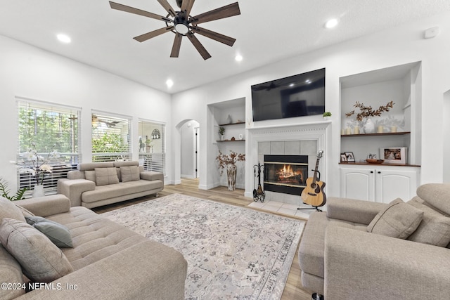 living room featuring built in shelves, ceiling fan, a fireplace, and light hardwood / wood-style floors