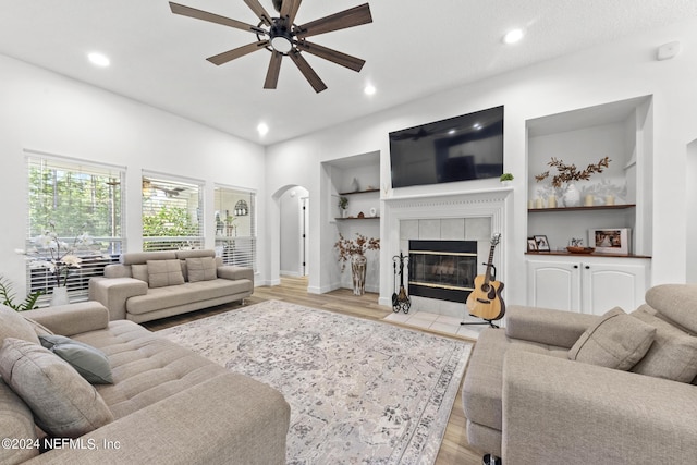 living room featuring built in shelves, ceiling fan, a tiled fireplace, and light wood-type flooring