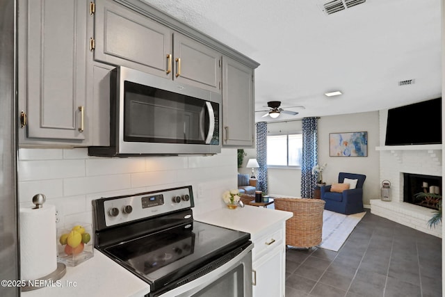 kitchen featuring gray cabinets, dark tile patterned floors, appliances with stainless steel finishes, backsplash, and a brick fireplace