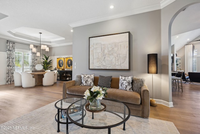 living room with arched walkways, ornamental molding, a tray ceiling, light wood finished floors, and an inviting chandelier