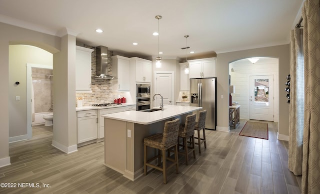 kitchen featuring white cabinetry, a center island with sink, pendant lighting, stainless steel appliances, and wall chimney range hood