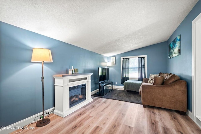 living room featuring a textured ceiling, light wood-style flooring, baseboards, vaulted ceiling, and a glass covered fireplace