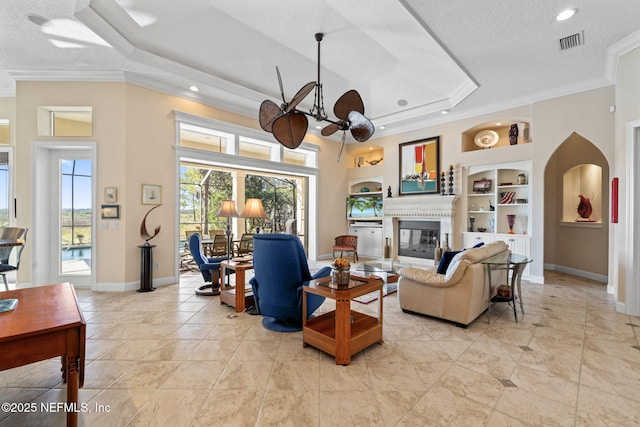 living room featuring visible vents, a glass covered fireplace, a tray ceiling, crown molding, and built in shelves