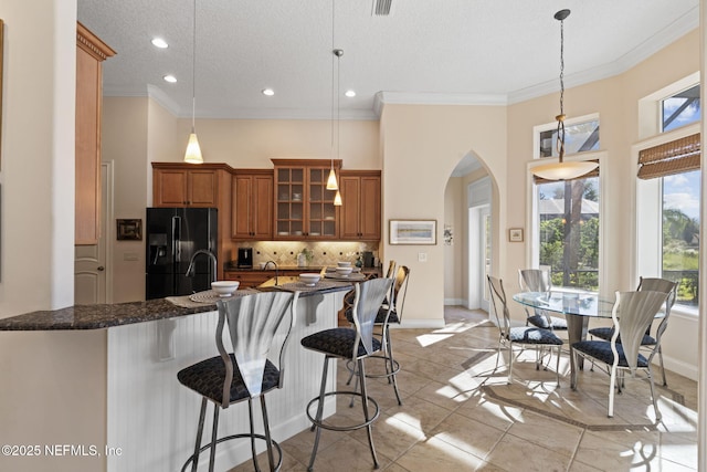 kitchen with a breakfast bar, black fridge with ice dispenser, decorative backsplash, brown cabinetry, and glass insert cabinets