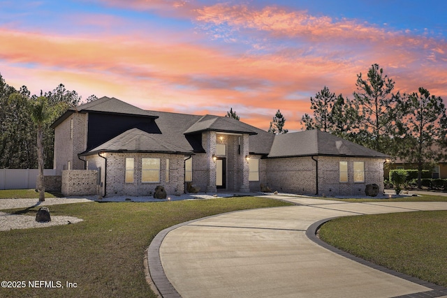 french country home featuring a shingled roof, brick siding, fence, a front lawn, and curved driveway