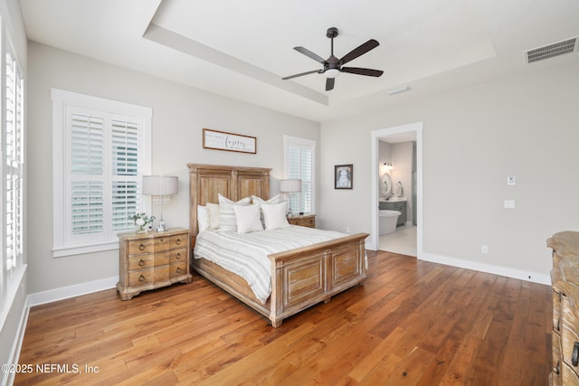 bedroom with ensuite bath, light hardwood / wood-style flooring, a raised ceiling, and ceiling fan