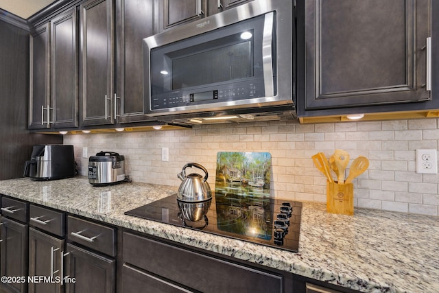 kitchen with black electric stovetop, stainless steel microwave, backsplash, dark brown cabinetry, and light stone countertops