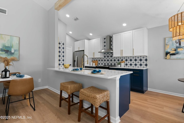 kitchen with blue cabinets, white cabinets, a kitchen bar, stainless steel refrigerator with ice dispenser, and wall chimney range hood