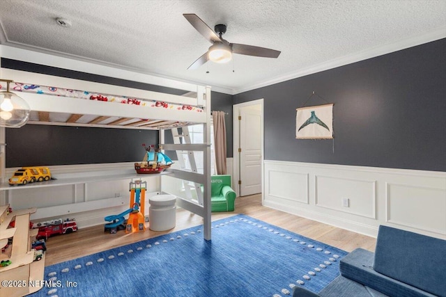 bedroom featuring a wainscoted wall, ornamental molding, a ceiling fan, a textured ceiling, and wood finished floors