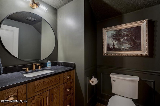 half bathroom with vanity, toilet, wainscoting, and a textured ceiling