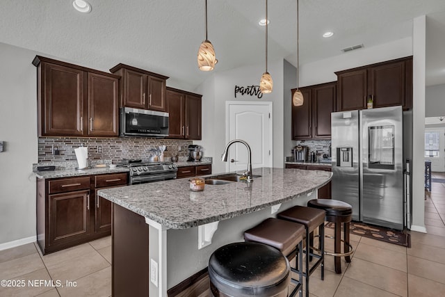 kitchen featuring pendant lighting, an island with sink, sink, stainless steel appliances, and light stone countertops