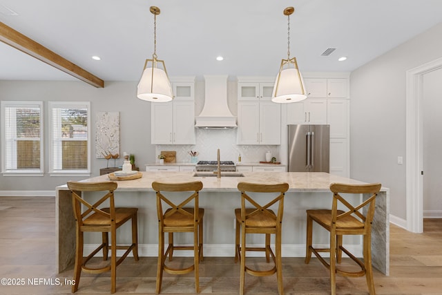 kitchen with custom exhaust hood, light stone countertops, high quality fridge, and a center island with sink