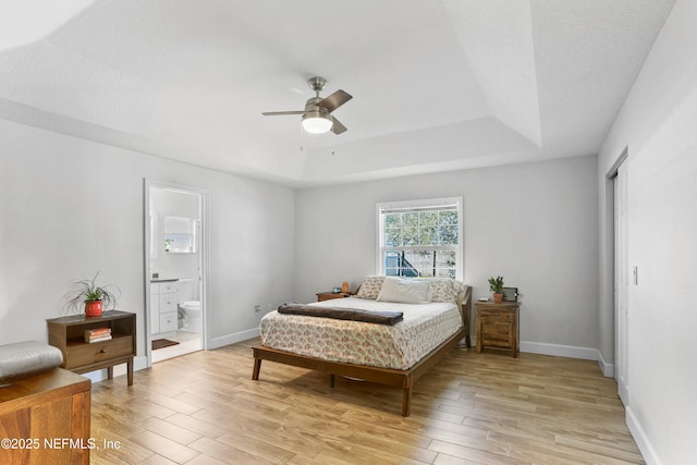 bedroom featuring light wood finished floors, baseboards, a ceiling fan, ensuite bathroom, and a tray ceiling