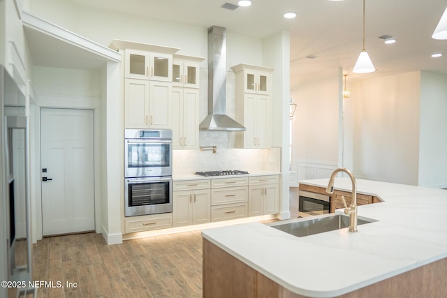 kitchen featuring wall chimney exhaust hood, sink, light hardwood / wood-style flooring, appliances with stainless steel finishes, and pendant lighting