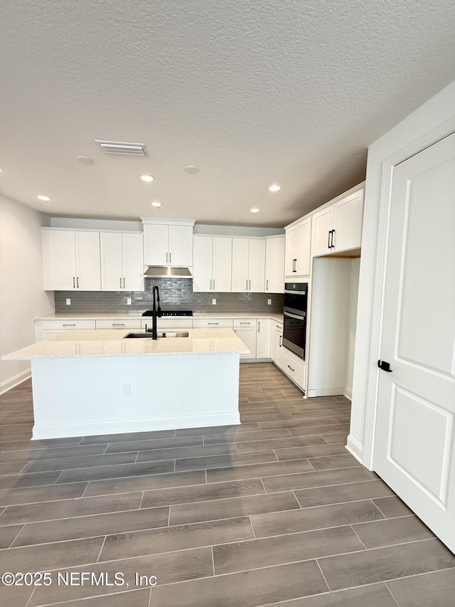 kitchen featuring double oven, white cabinetry, wood tiled floor, tasteful backsplash, and an island with sink