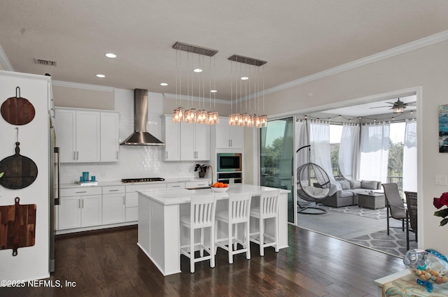 kitchen featuring white cabinetry, built in microwave, wall chimney range hood, hanging light fixtures, and an island with sink