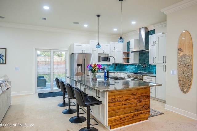 kitchen with dark stone counters, stainless steel appliances, wall chimney exhaust hood, hanging light fixtures, and a kitchen island with sink