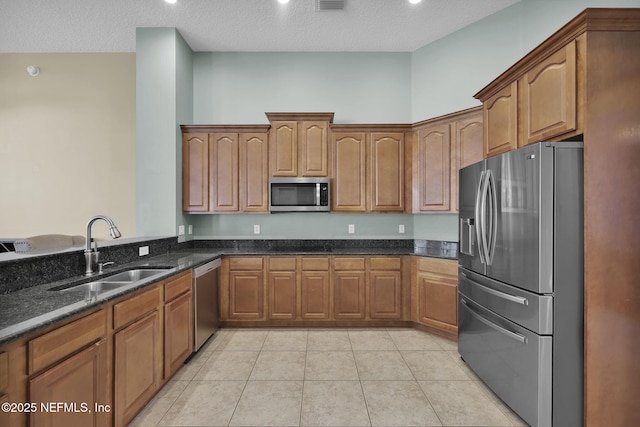 kitchen featuring light tile patterned floors, stainless steel appliances, brown cabinetry, a sink, and dark stone counters