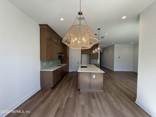 kitchen with decorative light fixtures, sink, dark hardwood / wood-style flooring, decorative backsplash, and a center island with sink