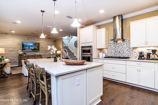 kitchen with an island with sink, white cabinetry, stainless steel appliances, hanging light fixtures, and wall chimney range hood