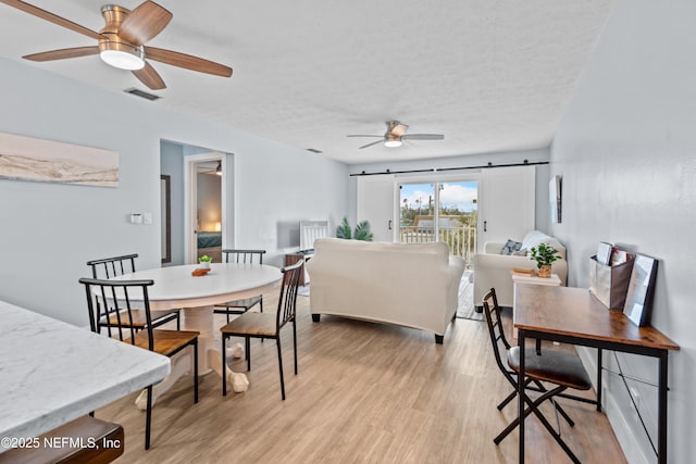 dining room with a textured ceiling, a barn door, light wood-style flooring, visible vents, and a ceiling fan