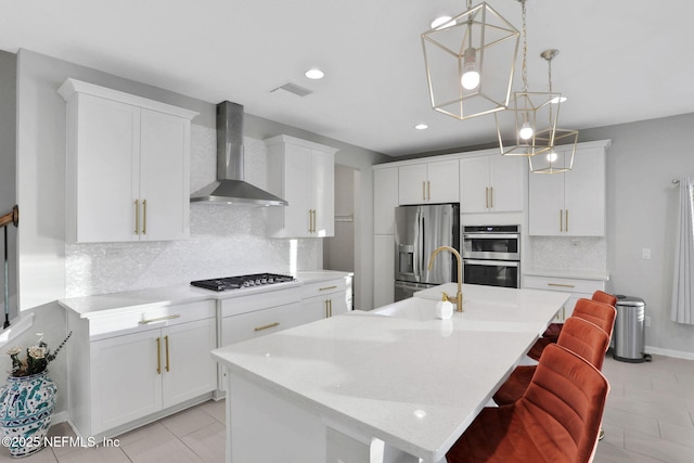 kitchen with a center island with sink, stainless steel appliances, light countertops, a sink, and wall chimney exhaust hood