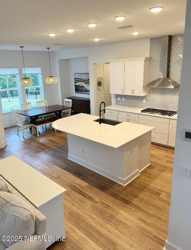 kitchen featuring a center island with sink, hanging light fixtures, wall chimney range hood, sink, and white cabinetry