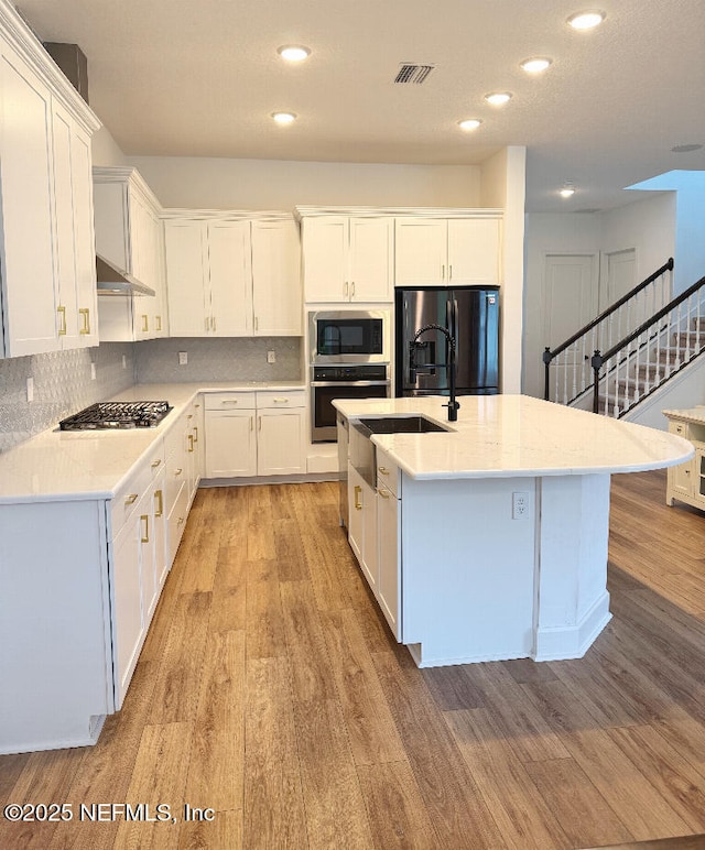 kitchen featuring appliances with stainless steel finishes, a center island with sink, light stone counters, light hardwood / wood-style floors, and white cabinetry