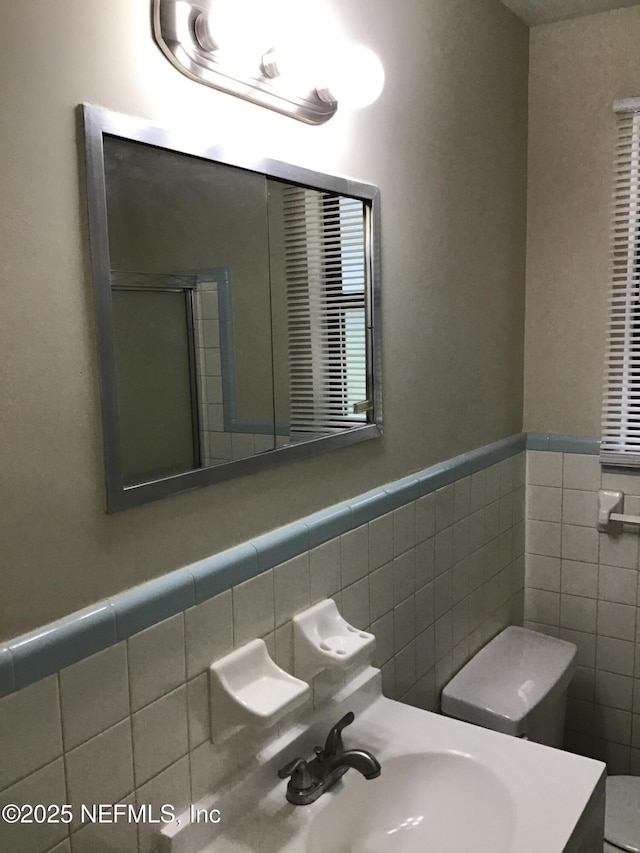 bathroom with tile walls, toilet, a stall shower, wainscoting, and a sink