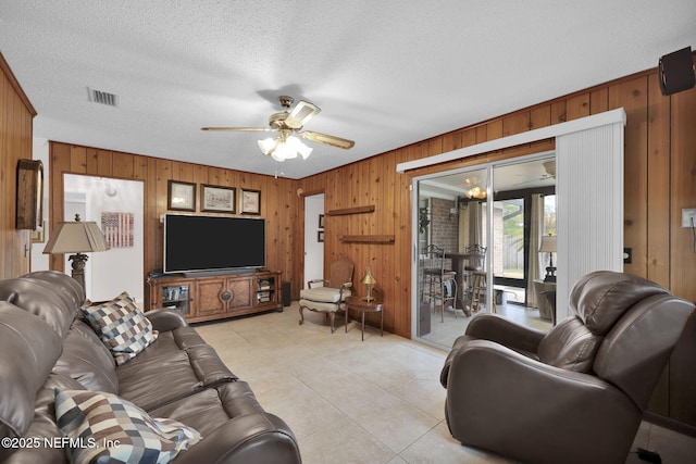 living area featuring a textured ceiling, light tile patterned floors, wood walls, visible vents, and a ceiling fan