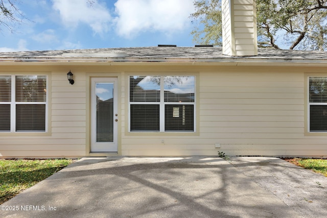 view of exterior entry with roof with shingles, a chimney, and a patio