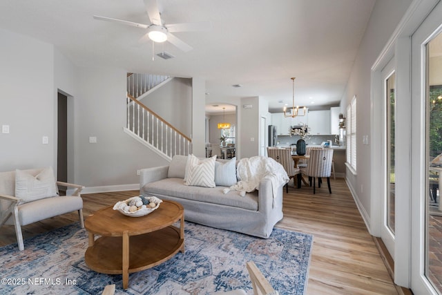 living area with stairs, visible vents, light wood-type flooring, baseboards, and ceiling fan with notable chandelier