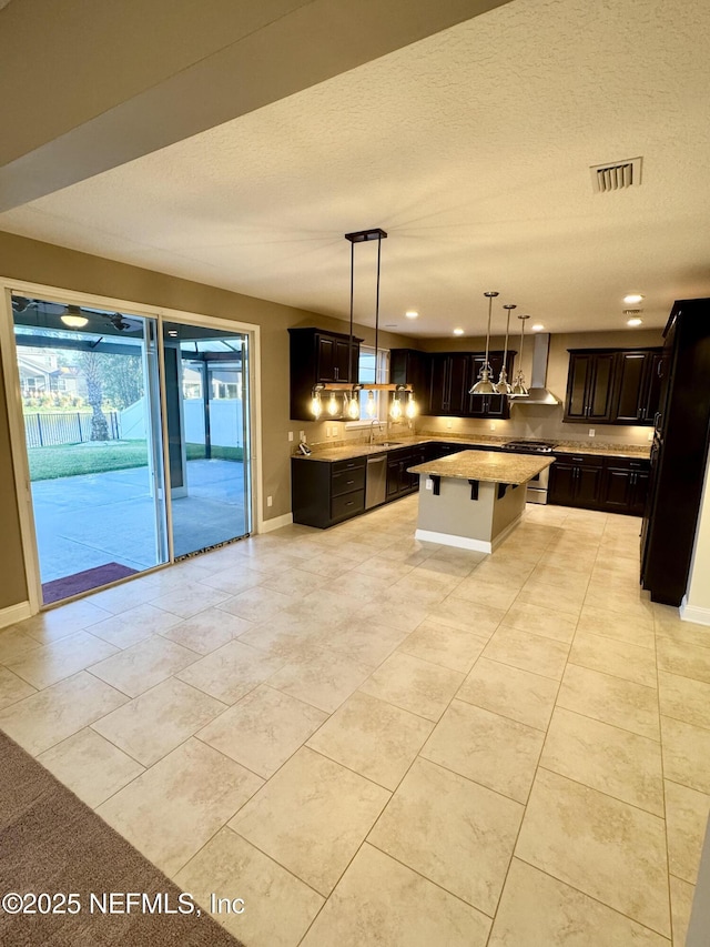 kitchen with dark brown cabinetry, a kitchen island, light countertops, wall chimney range hood, and decorative light fixtures