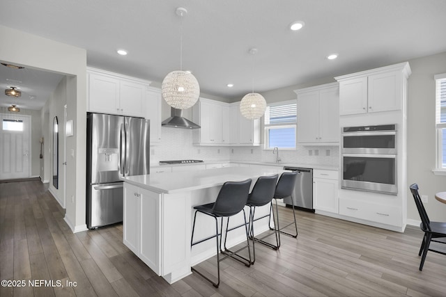 kitchen featuring a center island, appliances with stainless steel finishes, white cabinetry, a sink, and wall chimney range hood