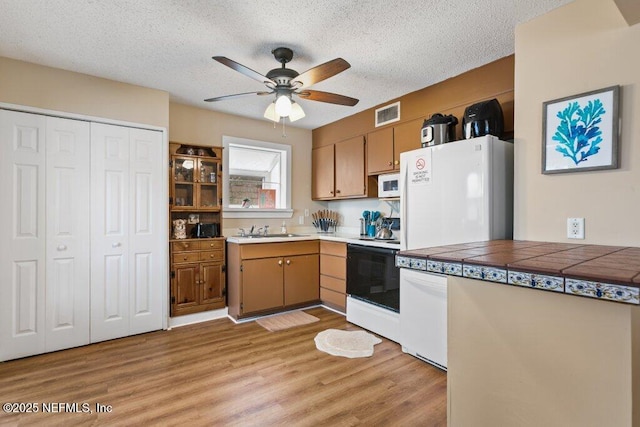 kitchen featuring tile countertops, brown cabinetry, a textured ceiling, light wood-type flooring, and white appliances