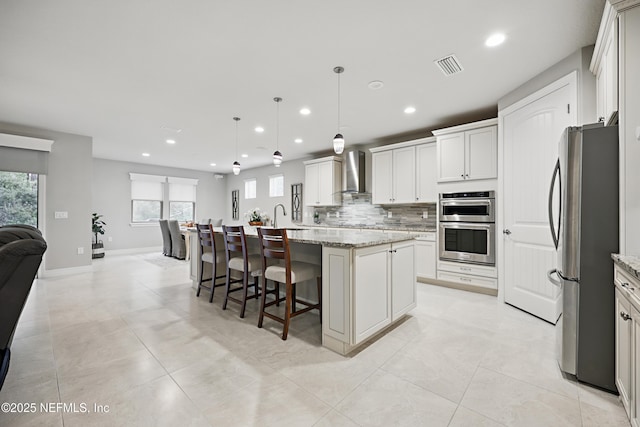 kitchen with light stone counters, stainless steel appliances, hanging light fixtures, wall chimney exhaust hood, and an island with sink