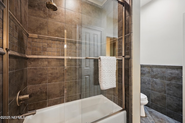 bathroom featuring toilet, bath / shower combo with glass door, tile walls, and wainscoting